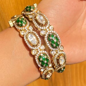 Puede incluir: Dos brazaletes dorados con detalles de esmeraldas verdes y piedras preciosas transparentes. Los brazaletes presentan piedras preciosas ovaladas y redondas engastadas en un marco dorado, con cierre de broche. El diseño incluye detalles intrincados y un acabado pulido.