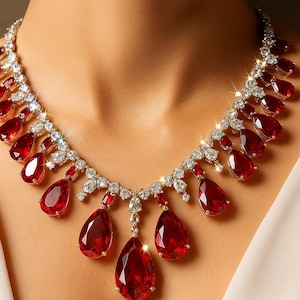 Könnte beinhalten: Eine atemberaubende Halskette mit großen, tropfenförmigen roten Edelsteinen und funkelnden, klaren Steinen. Die Halskette ist in einem silbernen oder weißen Metall gefasst und wird am Hals einer Person präsentiert.