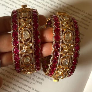 Könnte beinhalten: Zwei verzierte goldene Armreifen mit rubinroten Steinen und klaren, quadratisch geschliffenen Mittelsteinen. Die Armreifen weisen aufwendige Blumen- und geometrische Designs auf, mit kleinen, klaren Steinen für Glanz. Der Schmuck wird auf einer bedruckten Seite präsentiert.