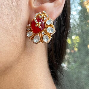 May include: A gold stud earring featuring a central oval red gemstone, encircled by clear, round gemstones and gold detailing. The design is elaborate and elegant, ideal for formal events or adding a touch of luxury.