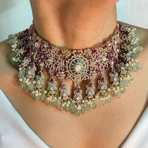 May include: A gold choker necklace with intricate floral designs, featuring pink and green gemstones. The necklace has a central pendant with a large white gemstone surrounded by smaller green gemstones. The necklace is adorned with dangling drops of green and white gemstones.