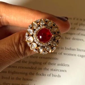 May include: A gold-toned ring with a large, round, red gemstone at the center. The ring is surrounded by smaller, clear gemstones and a decorative gold setting. The ring is held by a hand.