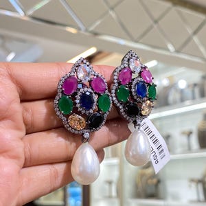 May include: A pair of ornate earrings with a cluster of colourful gemstones in shades of pink, green, blue, and yellow, surrounded by small clear stones. Each earring has a large, pear-shaped white pearl drop.