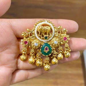 Puede incluir: Colgante dorado ornamentado con un motivo de elefante. El colgante presenta una gema verde central, rodeada de gemas más pequeñas y detalles dorados colgantes. El diseño incluye elementos florales y de cuentas.
