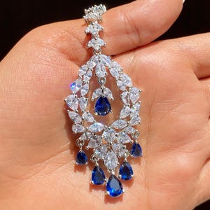 May include: Elegant chandelier earring featuring a teardrop design. The earring is adorned with clear, sparkling crystals and vibrant blue teardrop-shaped gemstones. A sophisticated piece of jewelry.