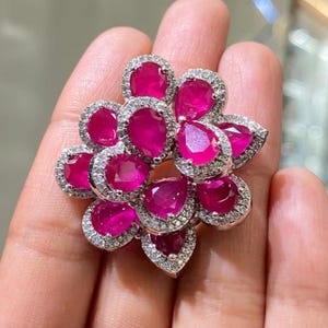 May include: A floral-shaped ring featuring vibrant, pear-cut ruby gemstones. Each ruby is surrounded by a halo of small, sparkling white diamonds, set in a silver-toned metal. The ring's design is intricate and eye-catching, with a focus on the rich color of the rubies.