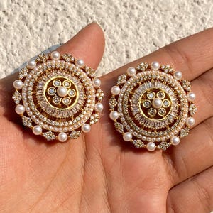 Pode incluir: Dois brincos dourados com um design circular. Cada brinco apresenta uma pérola central rodeada por pérolas menores, cristais transparentes e detalhes dourados. Os brincos têm uma estética vintage.