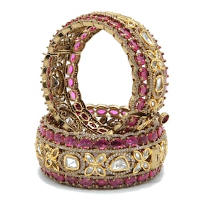 May include: Two gold-toned hinged bangles featuring rows of oval-shaped pink stones and clear, petal-shaped stones. The metal surface is encrusted with small, sparkling white crystals, creating a detailed, ornate texture across the entire band.