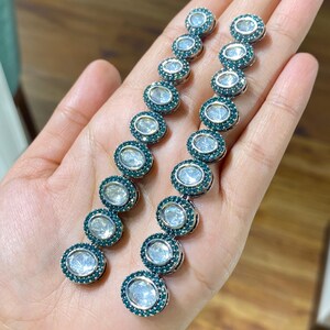 Pu&ograve; includere: Un paio di orecchini pendenti lunghi. Ogni orecchino presenta una serie di pietre ovali e trasparenti circondate da un anello di piccole pietre verde scuro. Gli orecchini sono tenuti in una mano, mostrando il loro design e colore.
