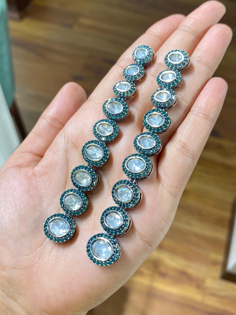 Pu&ograve; includere: Un paio di orecchini pendenti lunghi. Ogni orecchino presenta una serie di pietre ovali e trasparenti circondate da un anello di piccole pietre verde scuro. Gli orecchini sono tenuti in una mano, mostrando il loro design e colore.