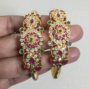 May include: Two gold bangles with detailed floral patterns. Each bangle showcases a central flower design with red and green gemstones, encircled by clear, sparkling stones. The bangles have a textured, coiled design near the opening.