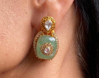 Könnte beinhalten: Goldfarbene Ohrringe mit einem hellgrünen, rechteckigen Mittelstein. Die Ohrringe haben ein Blumendesign in der Mitte und sind von kleinen, klaren Steinen umgeben. Oben befindet sich ein klarer, tropfenförmiger Stein.
