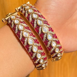 Pode incluir: Duas pulseiras douradas com base em esmalte vermelho, adornadas com pedras claras em forma de gota e pedras claras menores. As pulseiras apresentam um padrão floral.