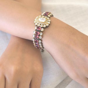 Pode incluir: Uma pulseira com uma pedra central retangular rodeada por pequenas pedras brancas, ladeada por filas de pedras ovais cor-de-rosa escuro cravadas em metal escuro. O design inclui detalhes intrincados em metal dourado à volta da pedra central.