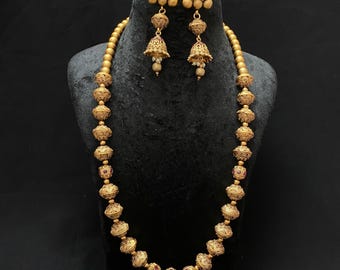 May include: A gold-toned necklace and earring set displayed on a black mannequin. The necklace features large, ornate beads interspersed with smaller, round beads. The earrings are bell-shaped with dangling accents. The jewelry set is likely made of metal.