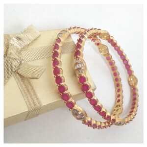 May include: Two gold-toned bangles featuring a continuous row of round, vibrant pink stones interspersed with small, clear, rectangular-cut stones. The jewelry is positioned next to a gold-colored gift box with a matching ribbon bow.
