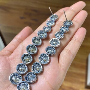 Pu&ograve; includere: Un paio di orecchini d'argento con una serie di pietre rotonde e trasparenti. Ogni pietra &egrave; incastonata in una lunetta d'argento con un bordo decorativo. Gli orecchini sono esposti su una mano.