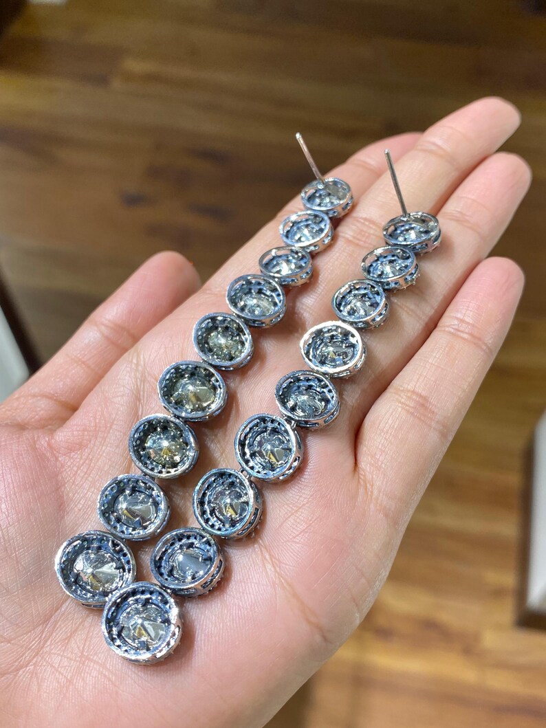 Pu&ograve; includere: Un paio di orecchini d'argento con una serie di pietre rotonde e trasparenti. Ogni pietra &egrave; incastonata in una lunetta d'argento con un bordo decorativo. Gli orecchini sono esposti su una mano.