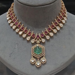 May include: A gold necklace with a large green gemstone pendant. The necklace is adorned with smaller white and red gemstones.