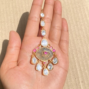 May include: A gold-coloured headpiece adorned with clear and pink gemstones. The piece showcases a central oval design with a pink gemstone, encircled by smaller pink stones and clear, teardrop-shaped gems. A chain of clear stones extends upwards.