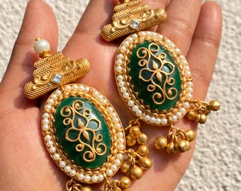 May include: Gold-toned earrings with a green oval center and a decorative gold design. The earrings are surrounded by small white pearls and have a cluster of gold beads hanging from the bottom. The top of the earrings have a small pearl and a square gem.