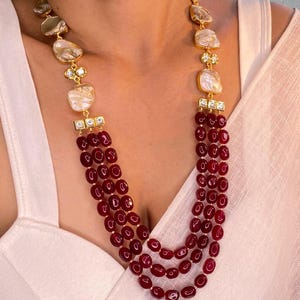 Puede incluir: Un collar de múltiples hilos con cuentas rojas y una cadena de oro con piedras blancas y doradas.