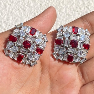 Puede incluir: Un par de pendientes cuadrados con una piedra roja redonda central rodeada por un grupo de piedras blancas claras rectangulares y en forma de pera, acentuadas con piedras rojas rectangulares adicionales en un marco de metal plateado.
