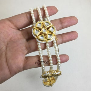 May include: An ornate, gold-toned bracelet with three strands of small, white pearls. The bracelet features a central floral design with mirrored accents and a white beaded border. The design is repeated at the bracelet's base. The bracelet is held in a hand.