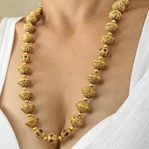 May include: A gold-toned necklace featuring large, ornate beads with intricate patterns. The necklace incorporates smaller beads and accents of red gemstones, creating a striking contrast. The design is symmetrical, with the beads evenly spaced along the chain.