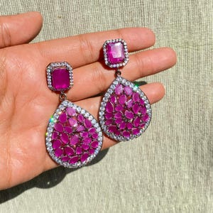 Peut inclure: Une paire de boucles d'oreilles pendantes magenta. Chaque boucle d'oreille présente une pierre carrée magenta en haut, reliée à un groupe en forme de goutte de petites pierres magenta, le tout encadré d'une bordure de petits cristaux transparents. Les boucles d'oreilles sont posées sur un fond neutre.