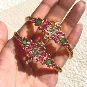 May include: Two gold-tone bangles featuring a central floral motif adorned with teardrop-shaped pink and green stones, accented by small clear round stones. The band of each bangle has a twisted, rope-like texture, creating a detailed design.