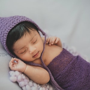 May include: A newborn baby wearing a purple crocheted hat and swaddled in a purple knitted wrap. The baby is sleeping peacefully on a white and purple textured surface.