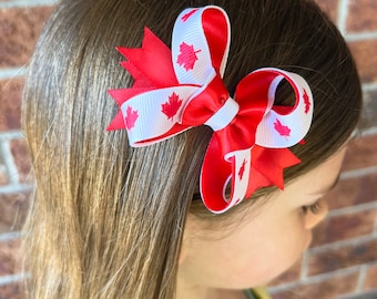 May include: A red and white bow with maple leaf designs. The bow is tied with a white ribbon and has a red centre.