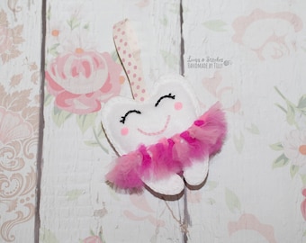 May include: A white felt tooth with a pink tulle tutu and a smiling face. The tooth is hanging from a pink and white polka dot ribbon. The text "Loops & Stitches Handmade by Tilly" is visible in the background.