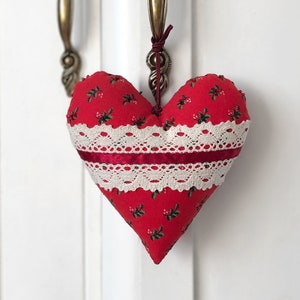 Pode incluir: Uma almofada vermelha em forma de coração com uma borda de renda branca e um pequeno padrão floral verde. A almofada está pendurada em uma maçaneta da porta.