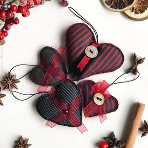 以下が含まれることがあります： 赤いリボンとボタンが付いた、黒と赤のストライプの布製のハートが3つ。ハートは、赤いベリー、シナモンスティック、スターアニスが置かれた白い背景に吊り下げられています。