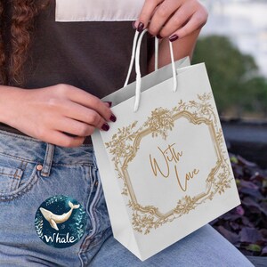 May include: A white gift bag with a gold floral design and the text "With Love" inside a frame. The bag is being held by a person wearing blue jeans.