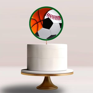 May include: A white cake on a gold cake stand, topped with a green-rimmed circle featuring a basketball, baseball, and soccer ball design. The cake is a dessert for a sports-themed event.