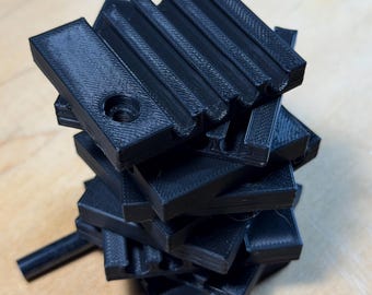 May include: A stack of black 3D-printed rectangular blocks. Each block has a series of parallel grooves on one side and a hole on the other. The blocks are arranged in a haphazard, slightly tilted tower.