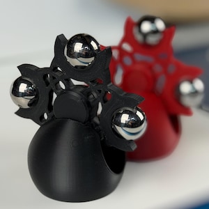 May include: Two fidget spinners, one black and one red, each with a unique design. The black spinner has a flower-like shape with four silver ball bearings. The red spinner is in the background. These are designed to be held in the hand.