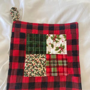 May include: A quilted Christmas potholder with a red and black buffalo plaid border. The center features four squares with holiday patterns, including holly and plaid designs. A small loop is attached for hanging.