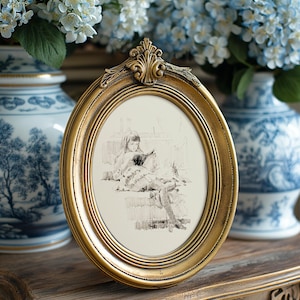 May include: An oval, gold-colored picture frame with an ornate top detail. The frame holds a black and white sketch of a person reading. Blue and white vases and flowers are in the background, suggesting a vintage or antique theme.