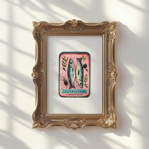 May include: A framed print of a sardine can illustration. The can is pink with two blue fish, olives, and sprigs of greenery. The text "Sardines A L'Huile D'Olive" is at the bottom. The frame is ornate gold.