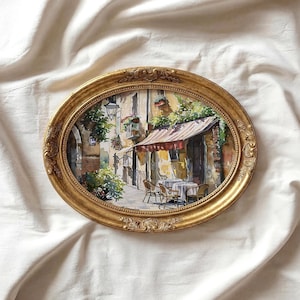 May include: An oval, gold-colored framed watercolor painting depicting a European street scene with a cafe. The painting features a red and white striped awning, tables, chairs, and lush greenery. The frame has ornate detailing.