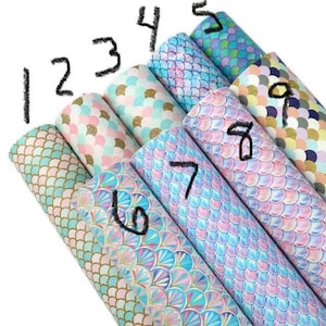 May include: A collection of nine rolls of faux leather fabric with mermaid scale patterns in various colors. The colors include shades of blue, pink, green, and gold. Each roll is numbered with a black marker.