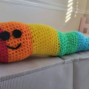 May include: A rainbow crocheted worm pillow with a smiling face. The pillow is made of red, orange, yellow, green, blue, and purple yarn.