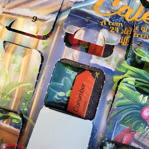 May include: An open advent calendar with several compartments visible. One compartment reveals a small packet labeled "Cucumber Cucumis" with an orange tag. Other compartments show images of plants and the numbers 2, 3, 9, 11, and 18.
