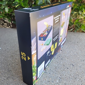 May include: A black box with gold text and illustrations. The box is for an Advent calendar, with the number 25 in gold on the side. The box has a QR code and images of the calendar's contents.