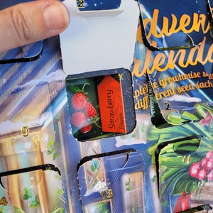 May include: An Advent calendar with a partially opened door revealing a small package labeled "Strawberry Fragaria" and a picture of strawberries. The calendar has a blue and gold design with numbers.