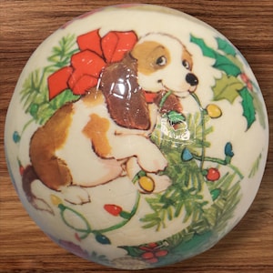May include: A round Christmas ornament featuring a cartoon puppy with brown and white fur, wearing a red bow. The ornament is decorated with a Christmas tree, holly, and colorful lights.